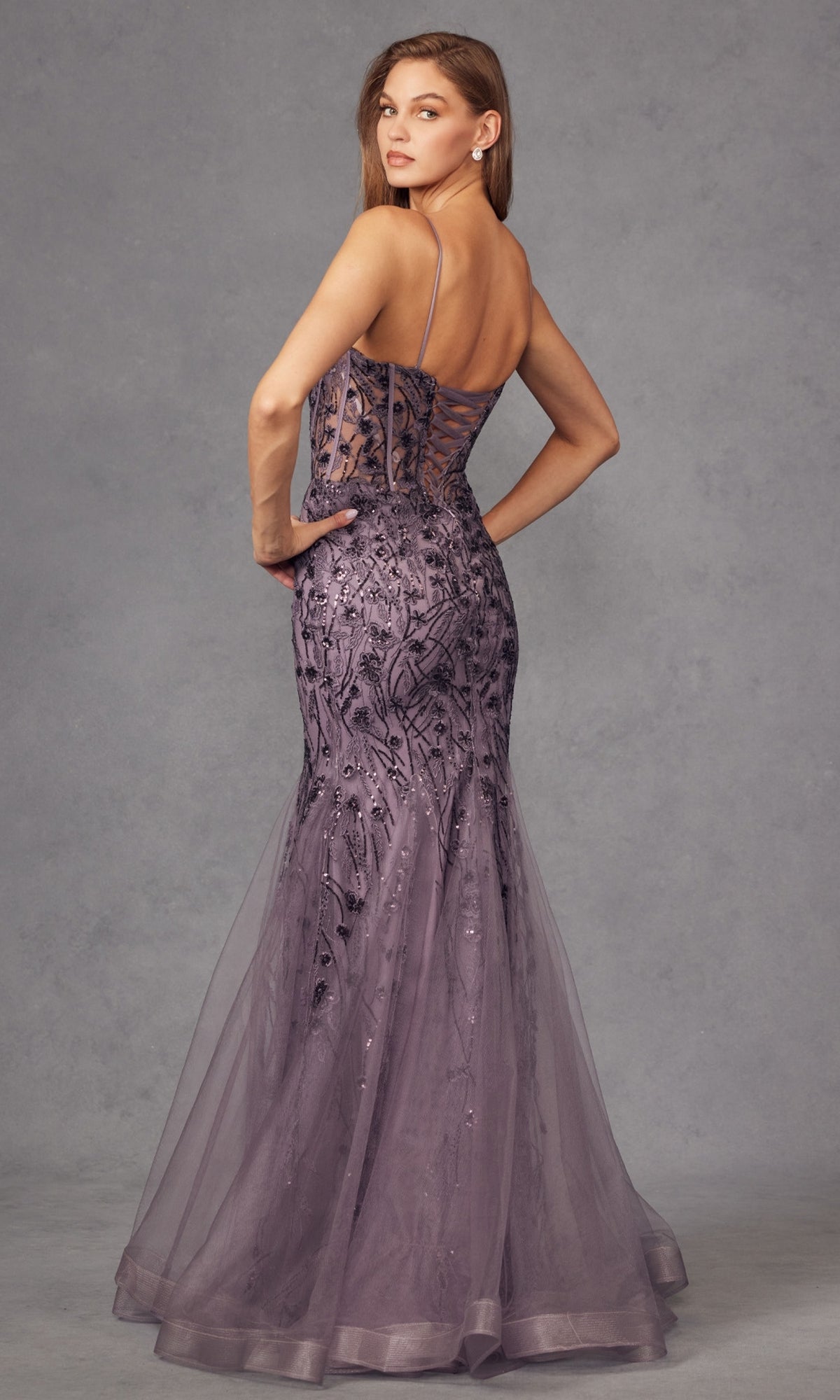 A woman poses in the Juliet JT2441S Long Sequin Mermaid Prom Dress, featuring a floor-length lavender design with thin straps, sheer panels, and intricate embroidery, set against a gray studio backdrop.